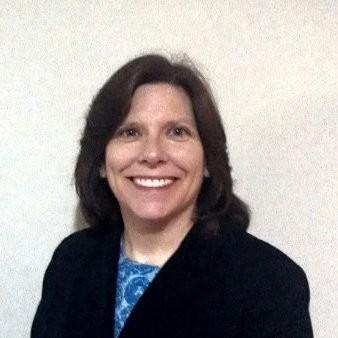 Smiling woman with shoulder-length brown hair, wearing a dark blazer over a blue patterned blouse, standing against a light wall.