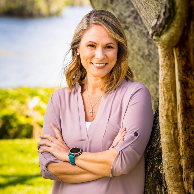 Smiling woman leaning against a tree outdoors, arms crossed, wearing a lavender blouse and cross necklace with a turquoise watch.
