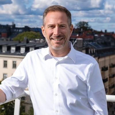 Smiling man in a white shirt standing on a balcony with a cityscape behind him.