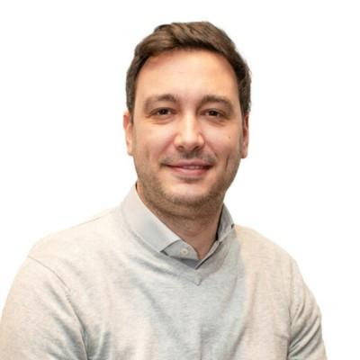 Head-and-shoulders portrait of a smiling man with short brown hair, wearing a light gray sweater, against a white background.