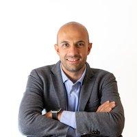 Portrait of a smiling bald man in a gray blazer and light blue shirt, with arms crossed against a white background.