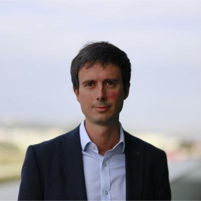 Portrait of a man in a navy blazer and light blue shirt, outdoors with a softly blurred urban background.
