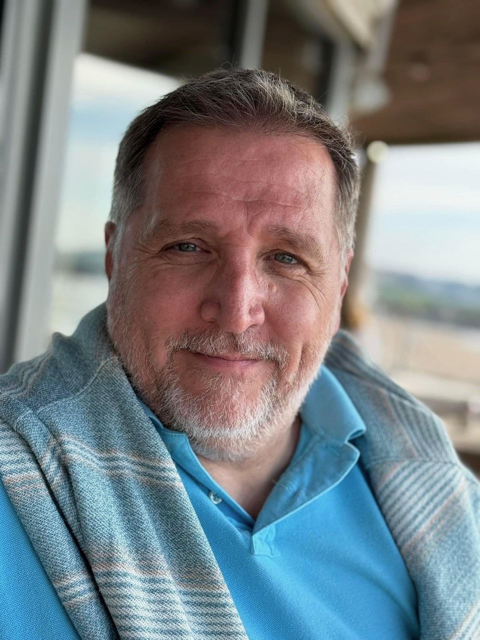 Smiling middle-aged man with short gray hair and beard, wearing a bright blue polo and a striped shawl, outdoors on a balcony.