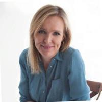 Smiling woman with shoulder-length blonde hair wearing a blue button-down shirt, seated against a white background.