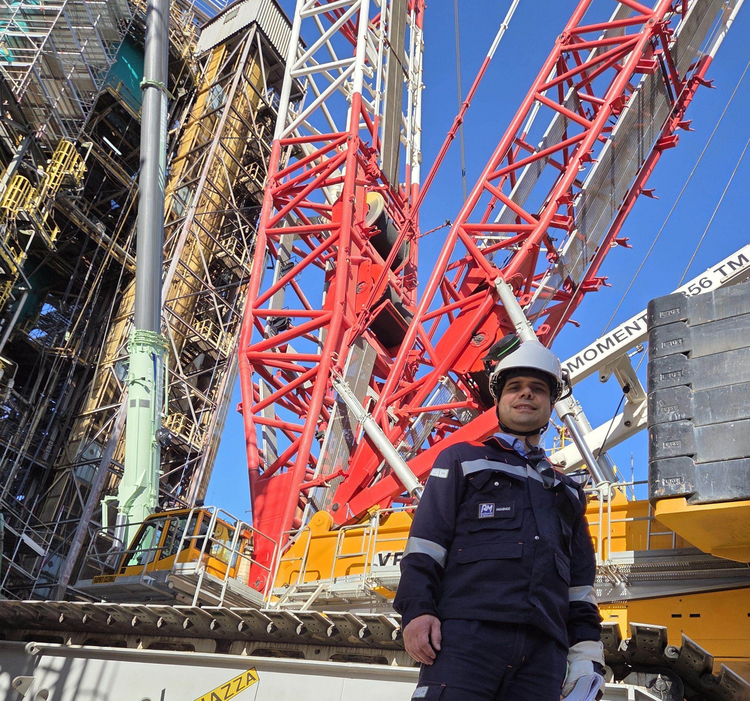 Construction worker in safety gear stands among red crane framework and dense scaffolding against a blue sky.