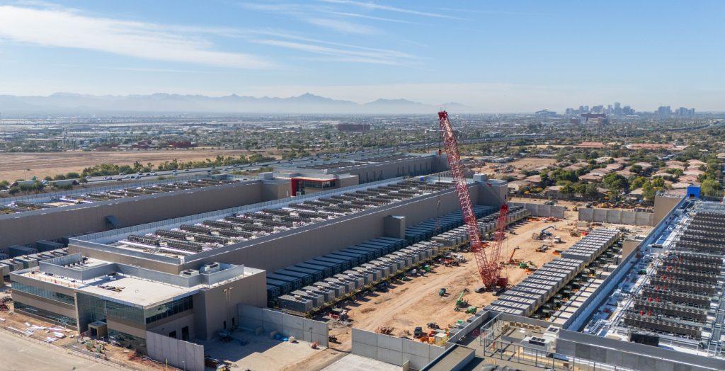 Aerial view of a large industrial construction site with a red crane long warehouse buildings and rows of equipment distant city skyline and mountains on the horizon