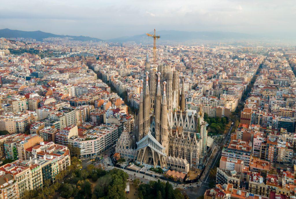 Aerial view of Barcelona with the Sagrada Família basilica in the foreground and a construction crane nearby, surrounded by dense city blocks and hills in the distance.