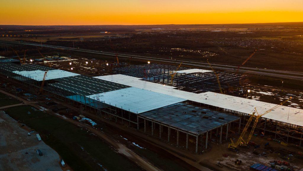 Aerial view of a large construction site at sunset with steel framework and several yellow cranes.