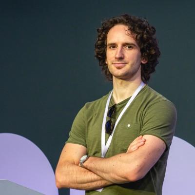 Smiling man with curly hair standing on a stage with arms crossed, wearing a green t-shirt and a conference lanyard.