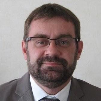 Portrait of a man with short dark hair, glasses, and a trimmed beard wearing a gray suit and tie.