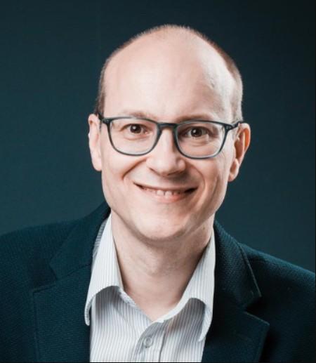 Smiling man with glasses in a head-and-shoulders studio portrait, wearing a blazer and light shirt.