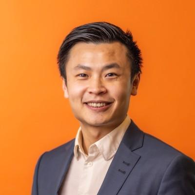 Head-and-shoulders portrait of a smiling Asian man in a navy blazer and light shirt against an orange background