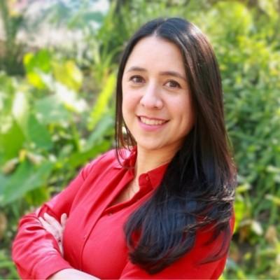 Smiling woman with long dark hair in a red blouse standing outdoors among greenery.