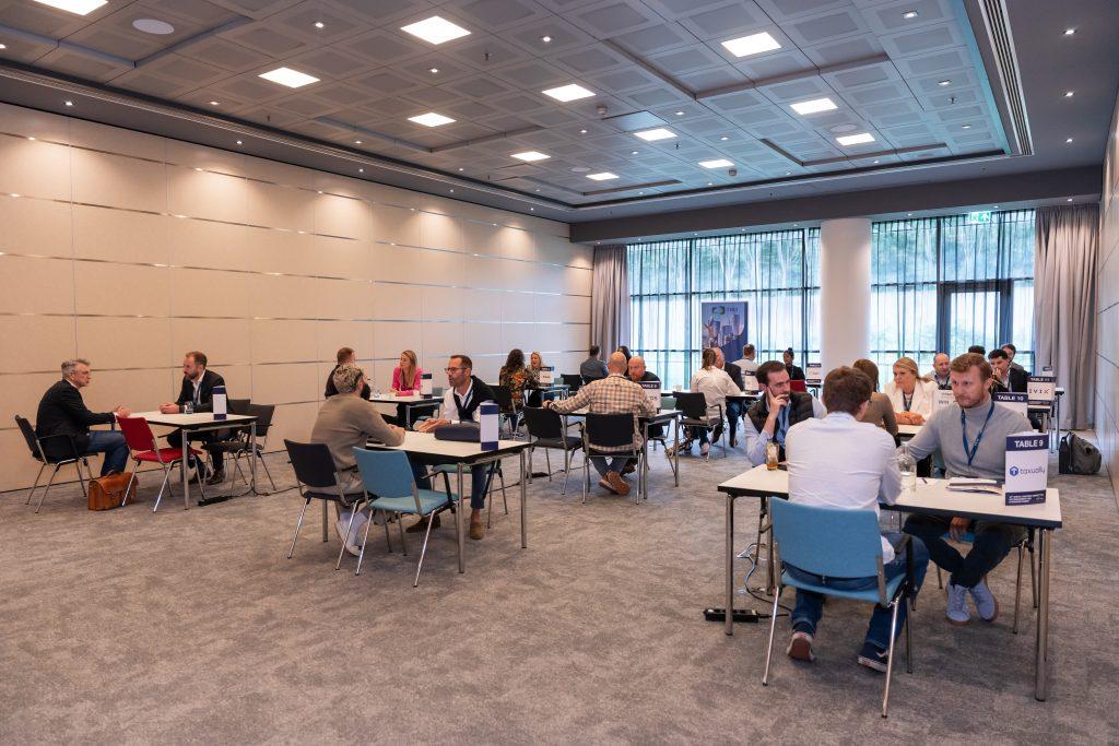 Conference hall during a networking event attendees sit at multiple tables and chat in small groups under bright ceiling lights