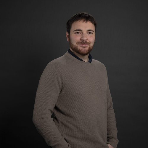 Portrait of a smiling man with short dark hair and a beard, wearing a beige sweater, against a dark studio backdrop.