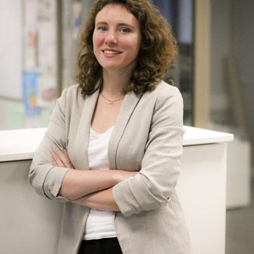 Smiling woman with curly brown hair wearing a light blazer, standing with arms crossed in an office setting.