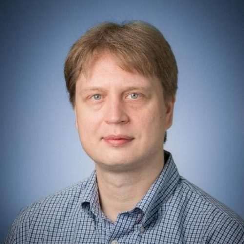 Head-and-shoulders portrait of a man with light brown hair in a blue checkered shirt against a blue gradient background.