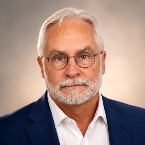 Head-and-shoulders portrait of an older man with white hair and beard, wearing round glasses and a navy suit, looking at the camera.