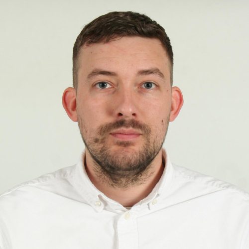 Close-up portrait of a man with short dark hair and stubble wearing a white button-down shirt, looking at the camera against a light background