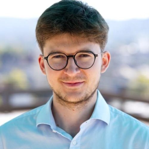 Portrait of a young man with round glasses and a light blue shirt outdoors on a sunny day, looking at the camera.