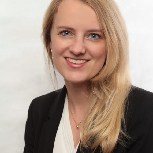 Portrait of a smiling blonde woman in a black blazer and white blouse against a light background.