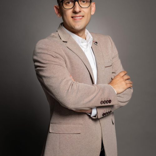 Portrait of a man wearing a beige blazer and white shirt, glasses, arms crossed, neutral studio backdrop.