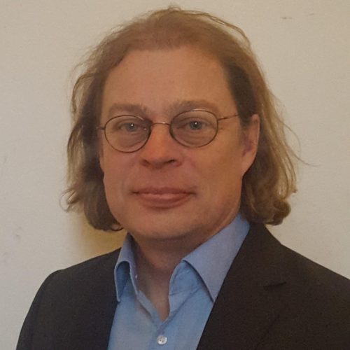 Portrait of a person in a blue shirt and dark blazer with round glasses and light brown hair, looking at the camera.