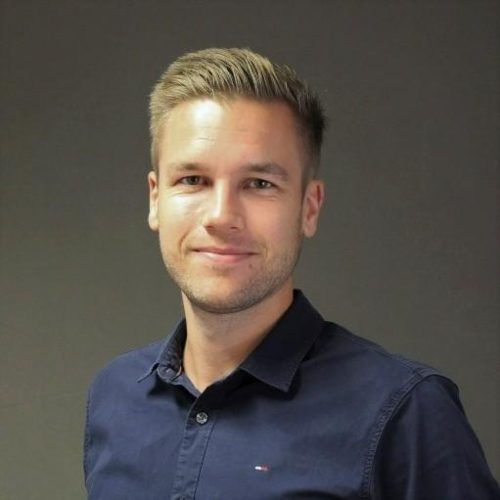 Smiling man in a dark blue button-down shirt posing for a head-and-shoulders portrait against a gray background.