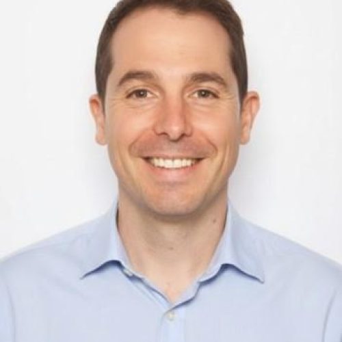 Smiling man in a light blue button-down shirt, facing the camera against a white background.