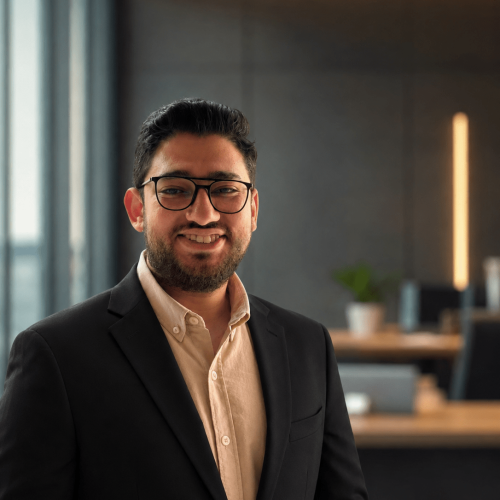 Portrait of a man with dark hair and glasses, wearing a beige button-up shirt and black blazer, smiling in a modern office setting.