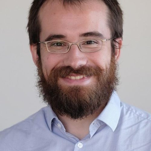 Smiling man with glasses and a full beard, wearing a light blue checkered shirt, looking at the camera.