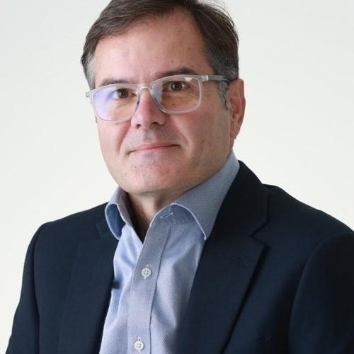 Portrait of a middle-aged man in a navy blazer and light blue shirt, wearing clear-framed glasses, facing the camera against a neutral background.