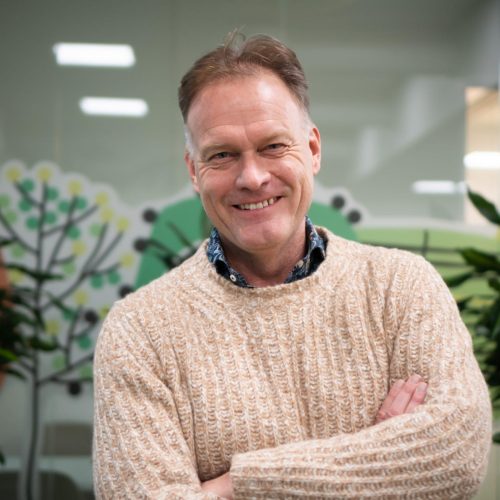 Smiling man in a cream knitted sweater stands with his arms crossed in a bright office, surrounded by plants and glass walls.