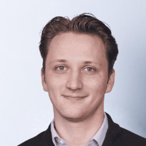 Head-and-shoulders portrait of a smiling man in a dark blazer and light shirt against a pale blue background in a professional headshot style.
