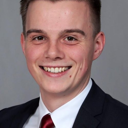 Head-and-shoulders portrait of a smiling man in a dark suit and red tie against a gray backdrop f.