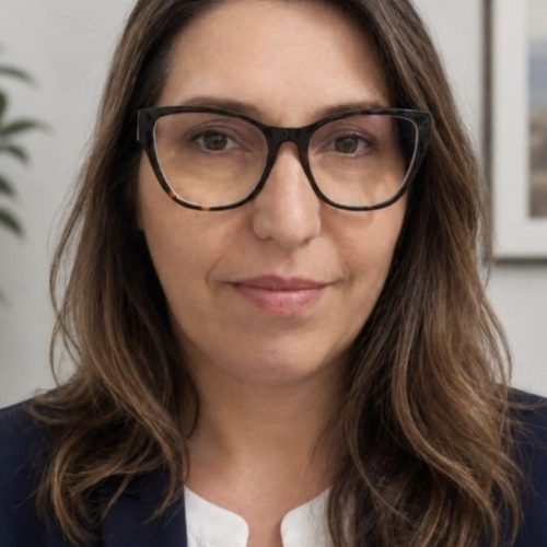 Portrait of a woman with shoulder-length brown hair and black-framed glasses, wearing a navy blazer and white blouse in a neutral room.