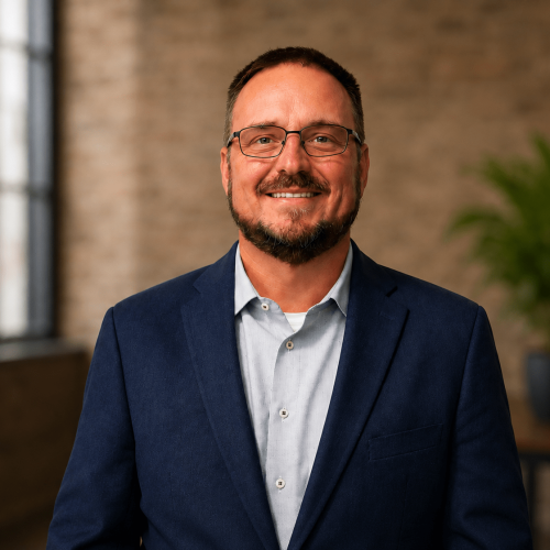 Smiling man in a navy blazer and light shirt, wearing glasses, in a brick-walled office with a plant in the background.