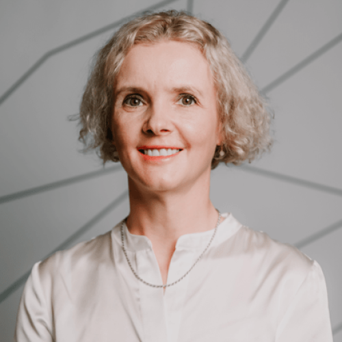 Portrait of a smiling woman with short gray hair wearing a white blouse, standing against a light gray geometric-patterned background.