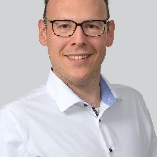 Professional headshot of a man wearing glasses and a white button-down shirt, smiling at the camera against a light gray background.
