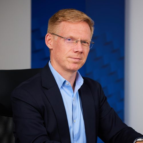 Portrait of a man in a navy blazer and light blue shirt wearing glasses, seated against a blue background.