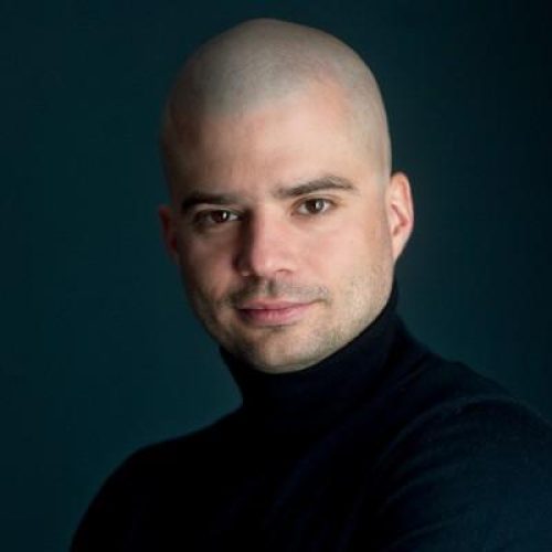 Head-and-shoulders portrait of a bald man in a black turtleneck, looking at the camera against a dark teal background.