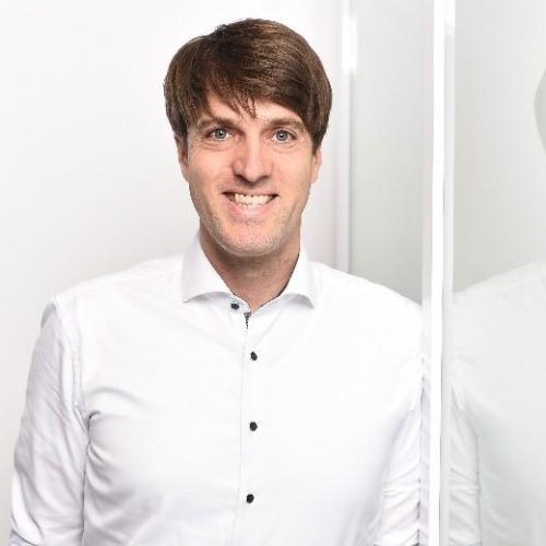 Smiling man in a white button-down shirt stands in a bright office beside a glass partition with his reflection visible in the glass, a professional headshot.