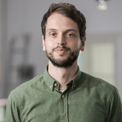 Portrait of a man with short brown hair and a beard wearing a green button-down shirt, standing in a bright office environment.