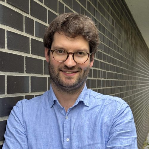 Smiling man with round glasses and a light beard stands in front of a dark brick wall wearing a blue button-up shirt.