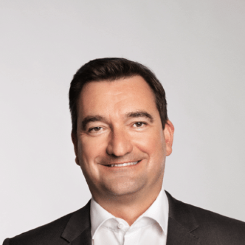 Head-and-shoulders portrait of a smiling man in a dark suit and white shirt, looking at the camera.