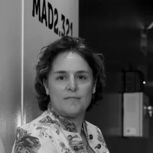 Portrait of a woman with short dark hair wearing a floral-patterned jacket, standing beside a wall with partial text visible (black-and-white photo).
