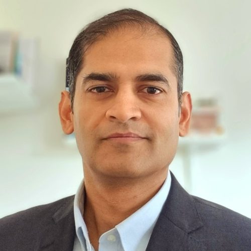 Portrait of a man in a dark blazer and light blue shirt looking at the camera in a neutral setting