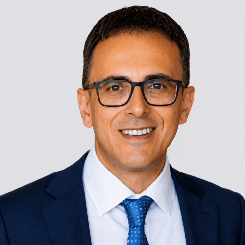Smiling man in a navy suit and blue tie wearing black glasses, portrait against a light gray background.