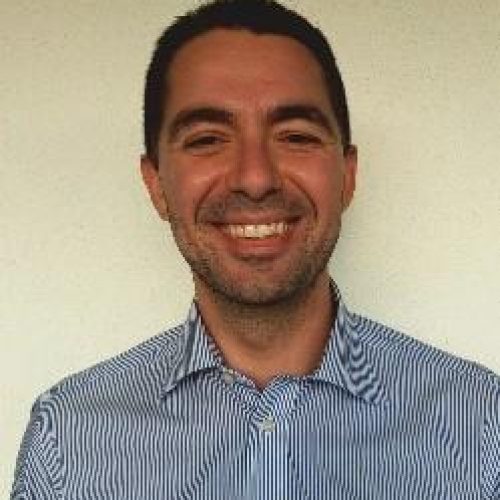 Head-and-shoulders portrait of a man in a light blue striped shirt smiling at the camera against a beige wall.