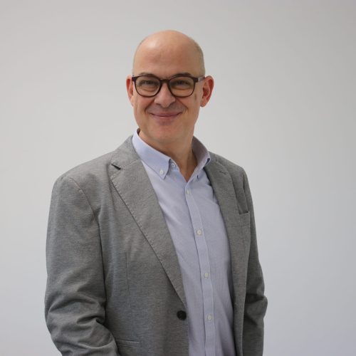 Smiling man in a grey blazer and light blue shirt wearing glasses, standing against a pale background (professional headshot).